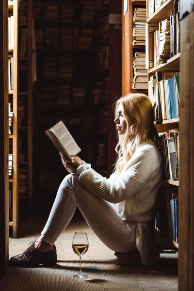 Vista laterale della donna bionda che legge il libro e siede sul pavimento in biblioteca — Foto stock
