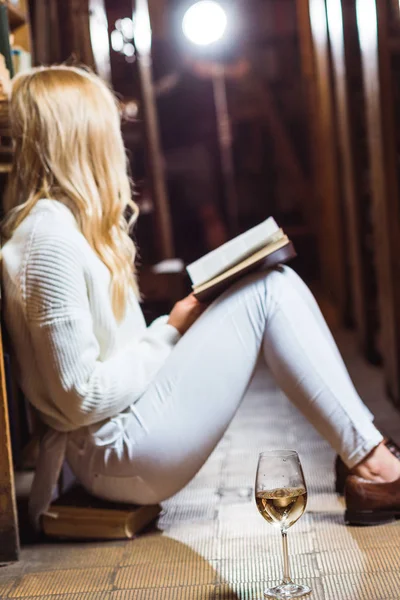Vista lateral da mulher loira lendo livro e sentado no chão na biblioteca — Fotografia de Stock