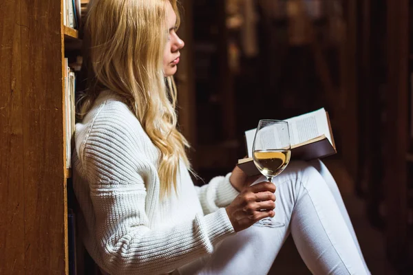 Vista lateral da mulher loira e bonita ler livro e segurando copo de vinho na biblioteca — Fotografia de Stock