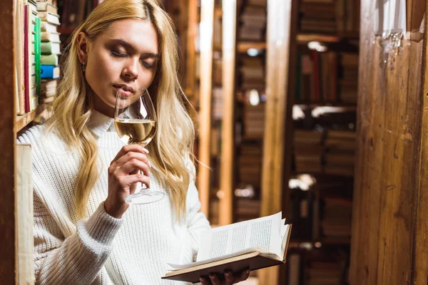 Femme blonde avec les yeux fermés tenant livre et verre à vin dans la bibliothèque — Photo de stock
