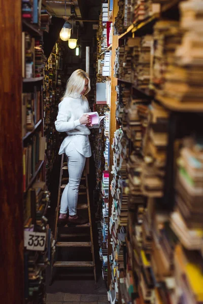 Donna bionda e carina in piedi sulla scala e libro di lettura in biblioteca — Foto stock
