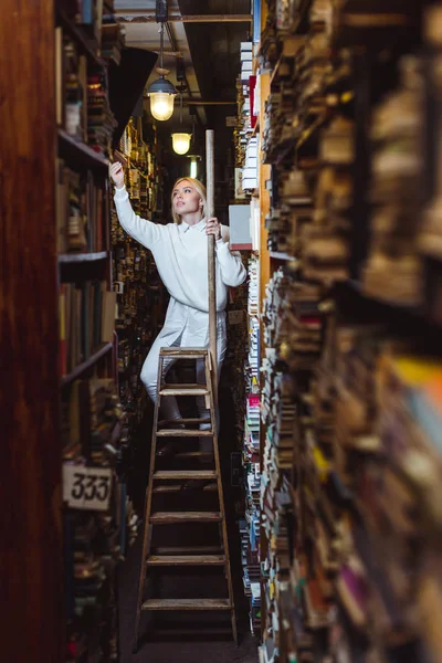 Loira e prettty mulher de pé na escada e segurando livro na biblioteca — Fotografia de Stock