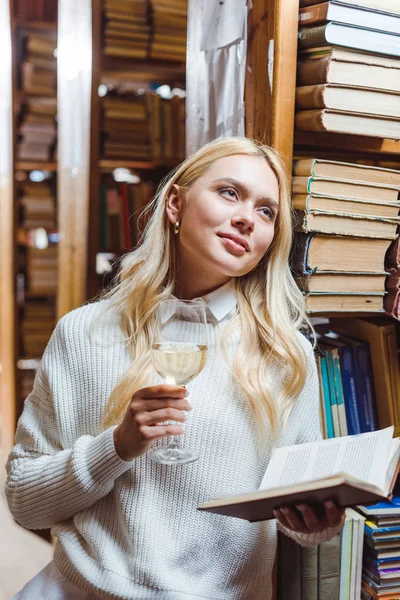 Mulher loira e bonita segurando livro e copo de vinho na biblioteca — Fotografia de Stock