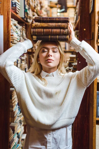 Rubia y bonita mujer en suéter blanco con los ojos cerrados sosteniendo libros en la biblioteca - foto de stock