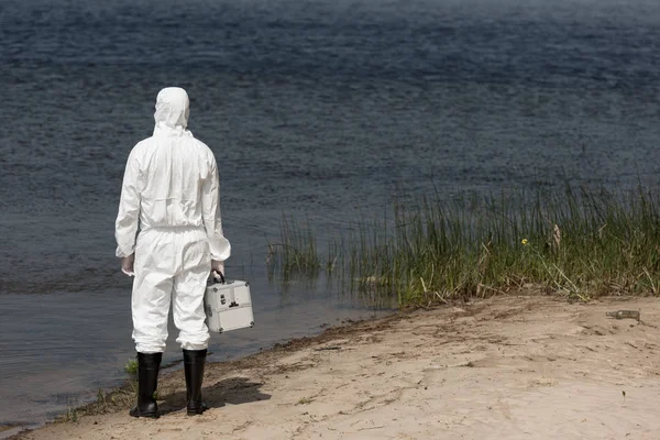 Back view of water inspector in protective suit holding inspection kit and standing on river coast — Stock Photo