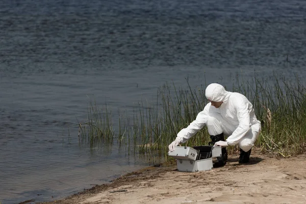 Inspector de agua en traje de protección con kit de inspección sentado en la costa del río - foto de stock