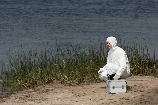 Inspector de agua en traje de protección con kit de inspección sentado en la costa del río - foto de stock