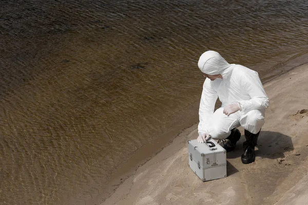 Water inspector in protective costume with inspection kit sitting on river coast — Stock Photo