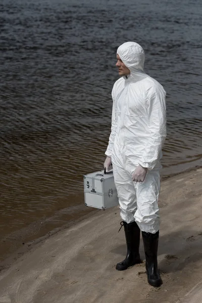 Full length view of water inspector in protective suit holding inspection kit and standing on river coast — Stock Photo