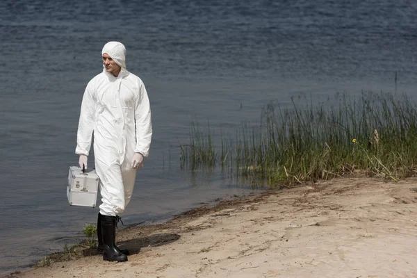 Full length view of water inspector in protective suit holding inspection kit and standing on river coast — Stock Photo