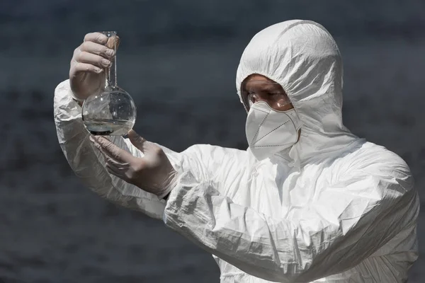 Inspetor de água em traje de proteção, respirador e óculos segurando frasco com amostra de água — Fotografia de Stock
