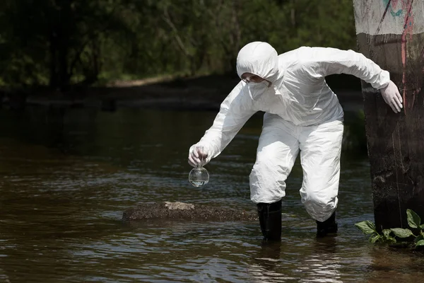 Inspecteur de l'eau en costume de protection, respirateur et lunettes tenant la fiole avec échantillon d'eau — Photo de stock