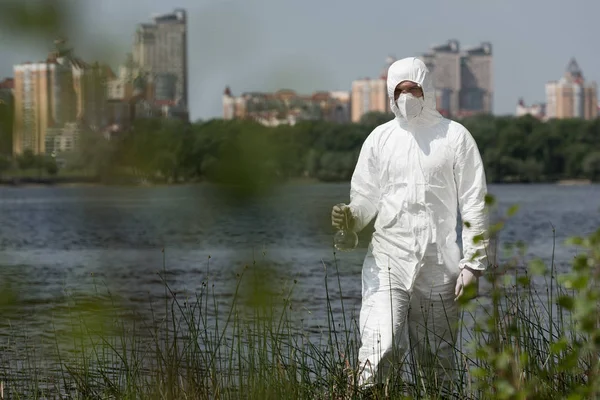 Selective focus of water inspector in protective costume, respirator and goggles holding flask with water sample — Stock Photo