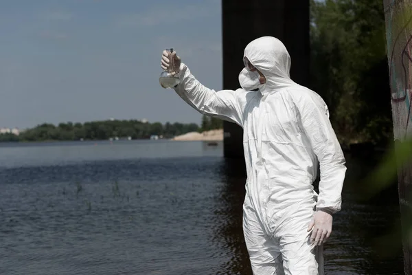 Water inspector in protective costume holding flask with water sample at river — Stock Photo