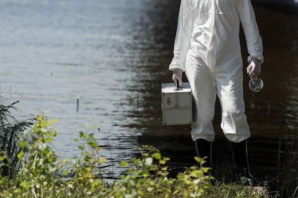 Partial view of water inspector in protective costume holding inspection kit and flask at river — Stock Photo