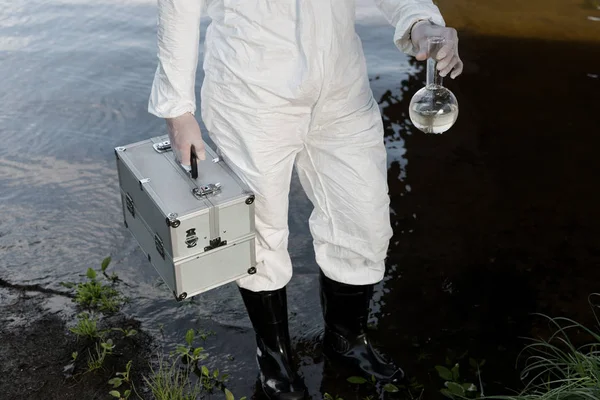 Partial view of water inspector in protective costume holding inspection kit and flask at river — Stock Photo