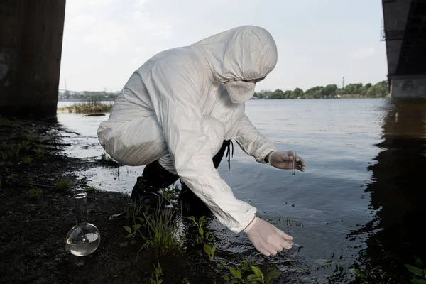 Water inspector in protective costume and respirator taking water sample at river — Stock Photo