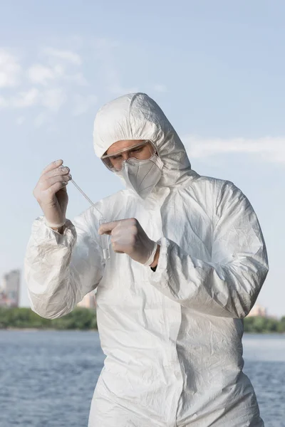 Water inspector in protective costume and respirator holding test tube with water sample at river — Stock Photo