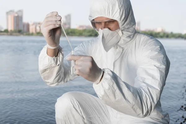 Wasserinspektor in Schutzkleidung und Atemschutzmaske entnimmt Wasserprobe am Fluss — Stockfoto