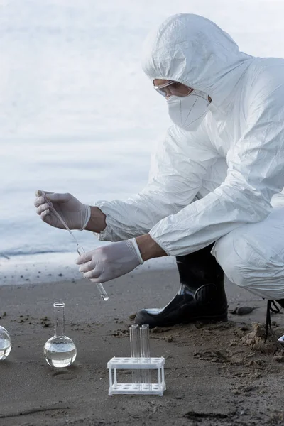 Wasserinspektor in Schutzkleidung und Atemschutzmaske entnimmt Wasserprobe am Fluss — Stockfoto