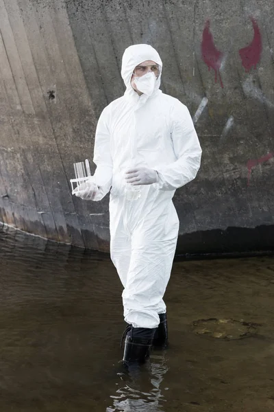 Full length view of water inspector in protective costume and respirator holding test tubes and flask at river — Stock Photo