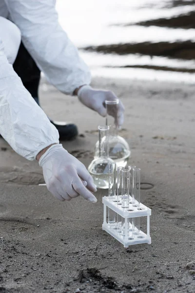 Cropped view of water inspector in latex gloves with test tubes and flasks at coast — Stock Photo
