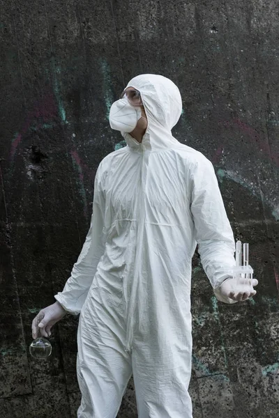 Water inspector in protective costume and respirator holding test tubes and flask with water samples at river — Stock Photo