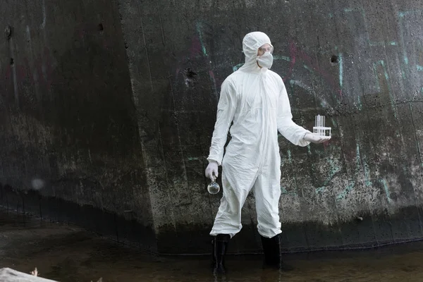 Full length view of water inspector in protective costume and respirator holding test tubes and flask with water samples at river — Stock Photo