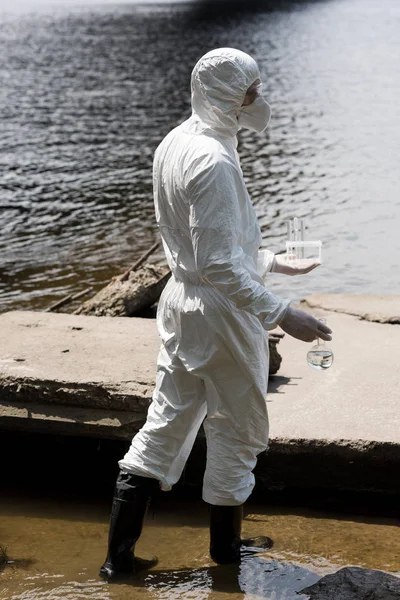 Side view of water inspector in protective costume and respirator holding test tubes and flask with water samples at river — Stock Photo