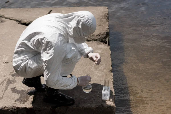 Wasserinspektor in Schutzkleidung, Latexhandschuhen und Atemschutzmaske entnimmt Wasserprobe am Fluss — Stockfoto