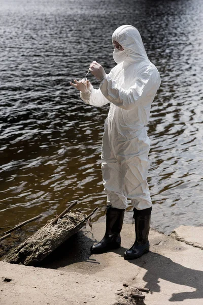 Ganzkörperansicht des Wasserinspektors in Schutzkleidung, Latexhandschuhen und Atemschutzmaske, der Wasserproben am Fluss entnimmt — Stockfoto