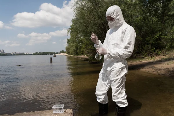 Water inspector in protective costume, latex gloves and respirator taking water sample at river — Stock Photo