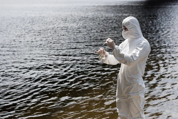 Water inspector in protective costume, latex gloves and respirator taking water sample at river — Stock Photo