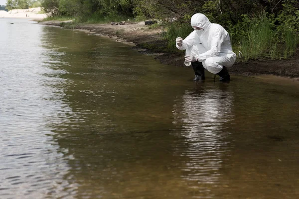 Инспектор по воде в защитном костюме, латексных перчатках и респираторе берет образец воды на реке — стоковое фото