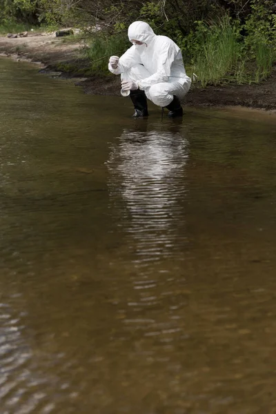 Inspecteur de l'eau en costume de protection, gants en latex et respirateur prélever un échantillon d'eau à la rivière — Photo de stock