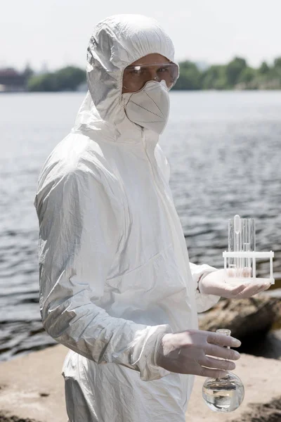 Water inspector in protective costume, latex gloves and respirator holding flask and test tubes with water samples at river — Stock Photo