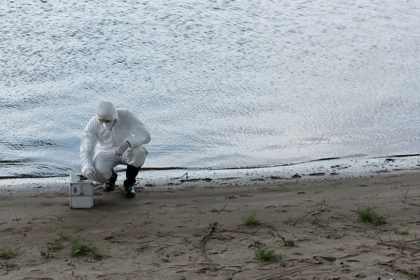 Water inspector in protective costume and respirator with inspection kit sitting at river coast — Stock Photo