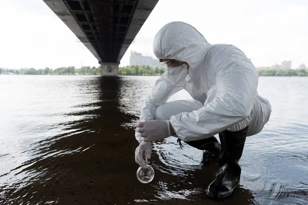 Water inspector in protective costume holding flask and taking water sample from river — Stock Photo