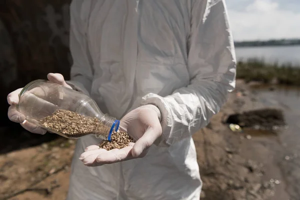 Cropped view of ecologist in protective costume holding bottle with pebbles — Stock Photo