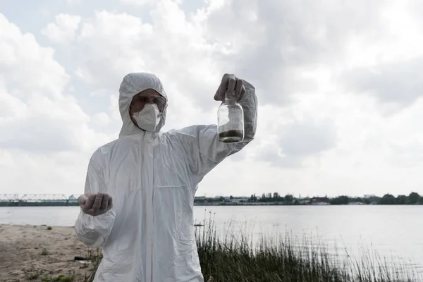 Ecologist in protective costume and respirator holding bottle with soil sample — Stock Photo