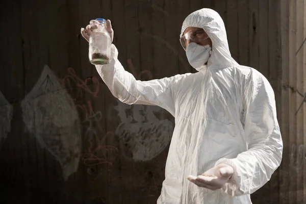Эколог в защитном костюме и респираторе держит бутылку с образцом почвы — стоковое фото