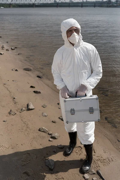 Full length view of water inspector in protective costume, respirator and goggles holding inspection kit on river coast — Stock Photo