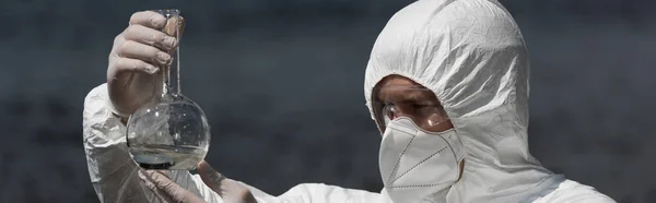 Panoramic shot of water inspector in respirator and goggles holding flask with water sample — Stock Photo