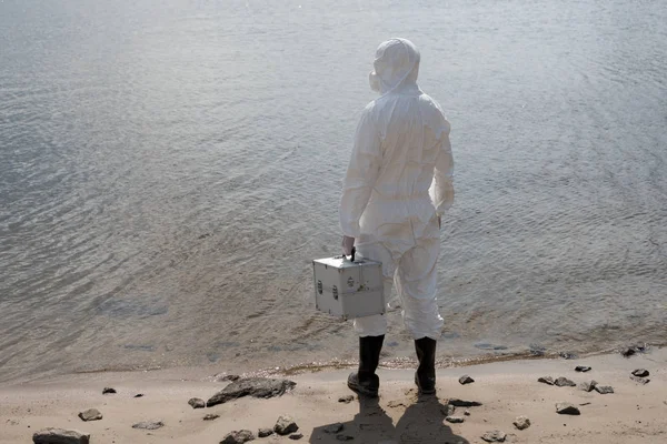 Back view of water inspector in protective costume holding inspection kit at river coast — Stock Photo