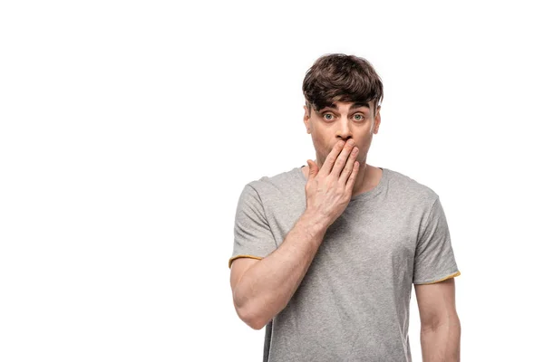 Shocked young man looking at camera while covering mouth with hand isolated on white — Stock Photo