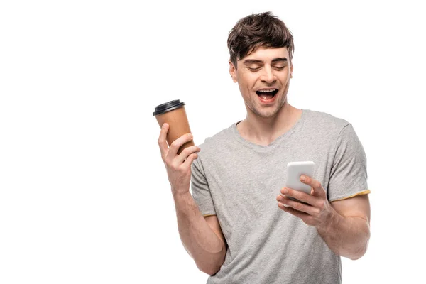 Alegre joven sosteniendo café para llevar mientras se utiliza el teléfono inteligente aislado en blanco - foto de stock