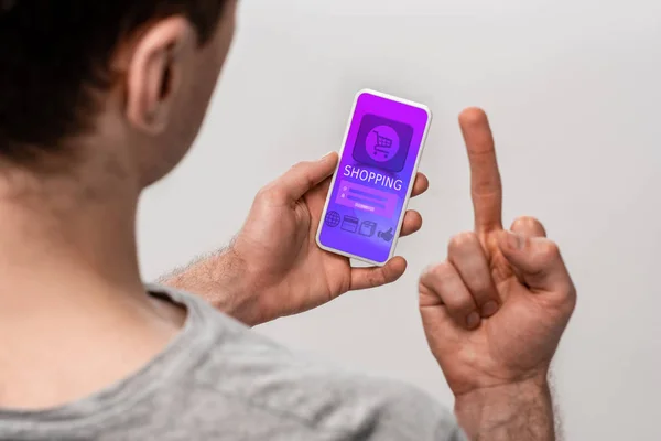 Vista recortada del hombre usando el teléfono inteligente con la aplicación de compras y mostrando el dedo medio, aislado en gris - foto de stock