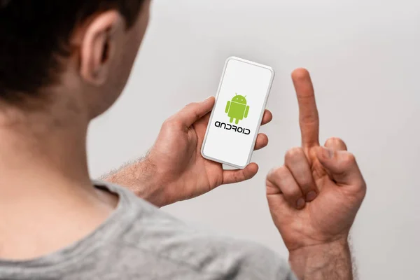 КИЕВ, УКРАИНА - 16 мая 2019 года: обрезанный вид человека с помощью смартфона с приложением для Android и показывающий средний палец, изолированный на сером — стоковое фото