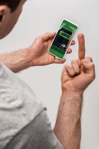 Vista recortada del hombre utilizando el teléfono inteligente con aplicación de reserva y mostrando el dedo medio, aislado en gris - foto de stock