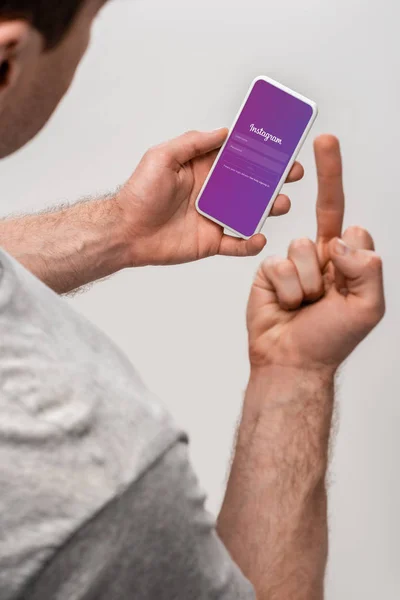 KYIV, UKRAINE - MAY 16, 2019: cropped view of man using smartphone with instagram app and showing middle finger, isolated on grey — Stock Photo